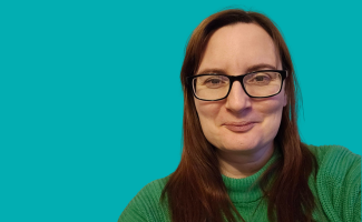 Headshot of a woman smiling. She has long dark brown hair and is wearing glasses. She is wearing a green jumper.