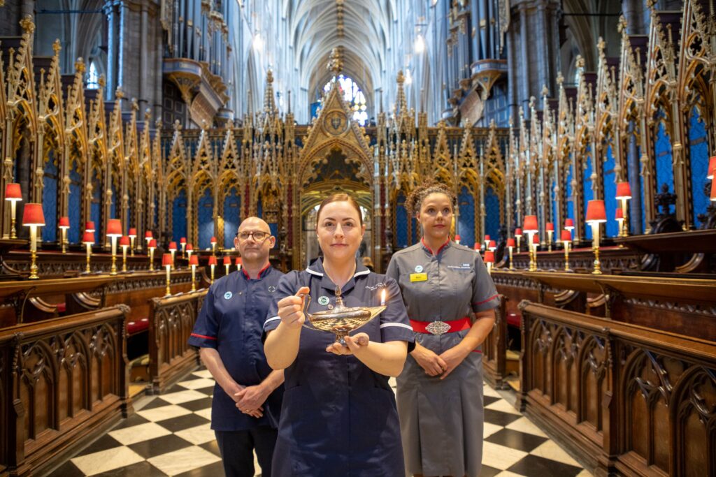 Inside Westminster Abbey, three people are standing, the middle person in holding the lamp used at the Florence Nightignale Commemoration Service, they are all in nursing/midwifery uniform.