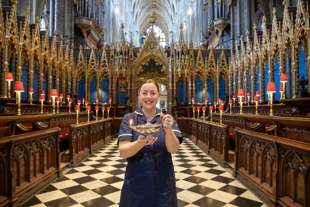 Woman standing on the black and white floor in Westminster Abbey, wearing a health care uniform, which is drak blue. She is holding a lamp to represent Florence Nightingales lamo. and she is smiling.