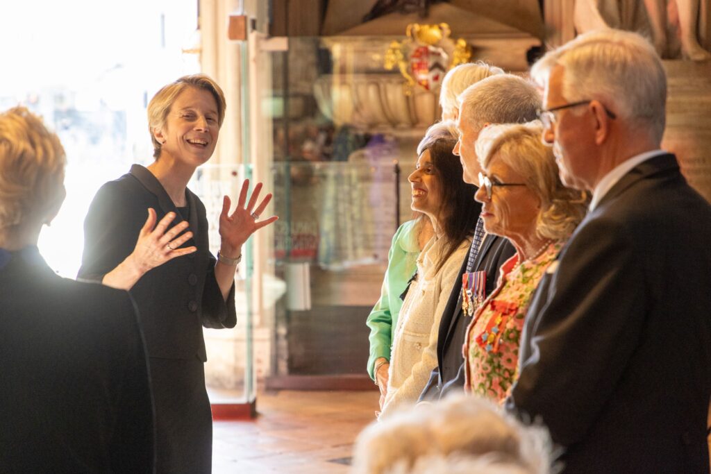 A woman in black on the left side of the image speaking to a group of people listening. She has had fingers and hands raised for emphasis as she speaks.