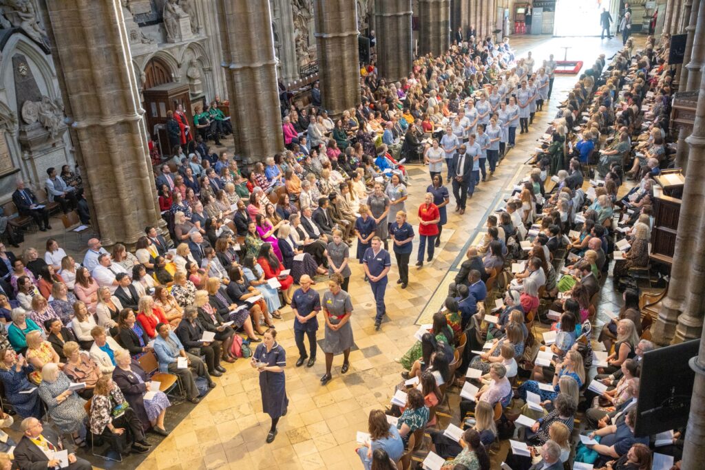 The inside of Westminster Abbey with a procession of nurses and midwives walking and people seated in rows on each side watching.