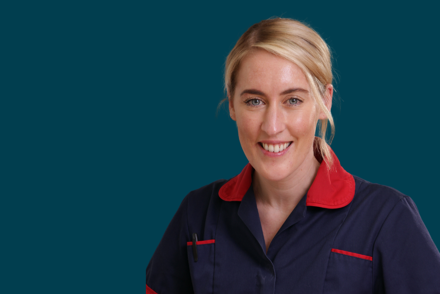 Head and shoulders image of a woman in navy and red nursing uniform. She has tied back blonde hair and is smiling.