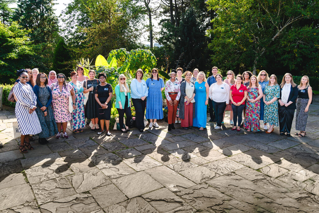 A group of people standing in a line smiling. They are outside and the sun is shining and there are trees and foliage in the background.