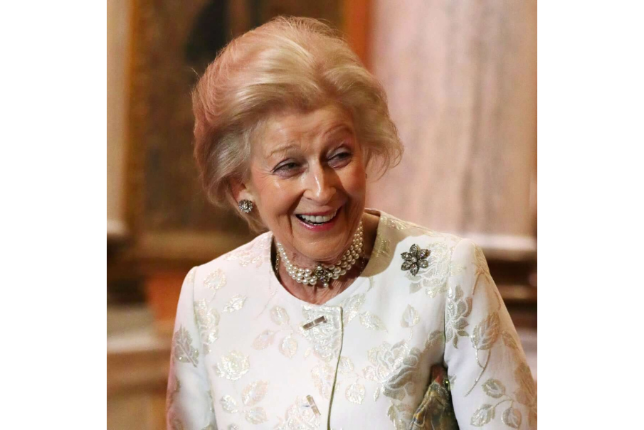 Head and shoulders image of a woman with blode hair which is chin length and swept back, she is looking to the right of the frame and smiling. She is wearing a cream jacket with a brooch on the shoulder and a pearl necklace.