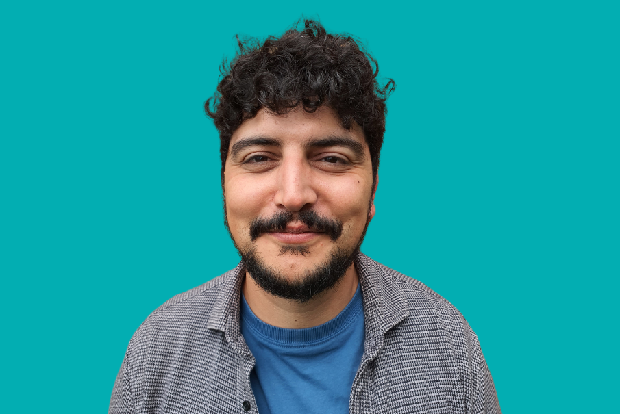 Headshot of a man with dark curly hair, and a beard and moustache, smiling. Hi is wearing a shirt with a t-shirt.