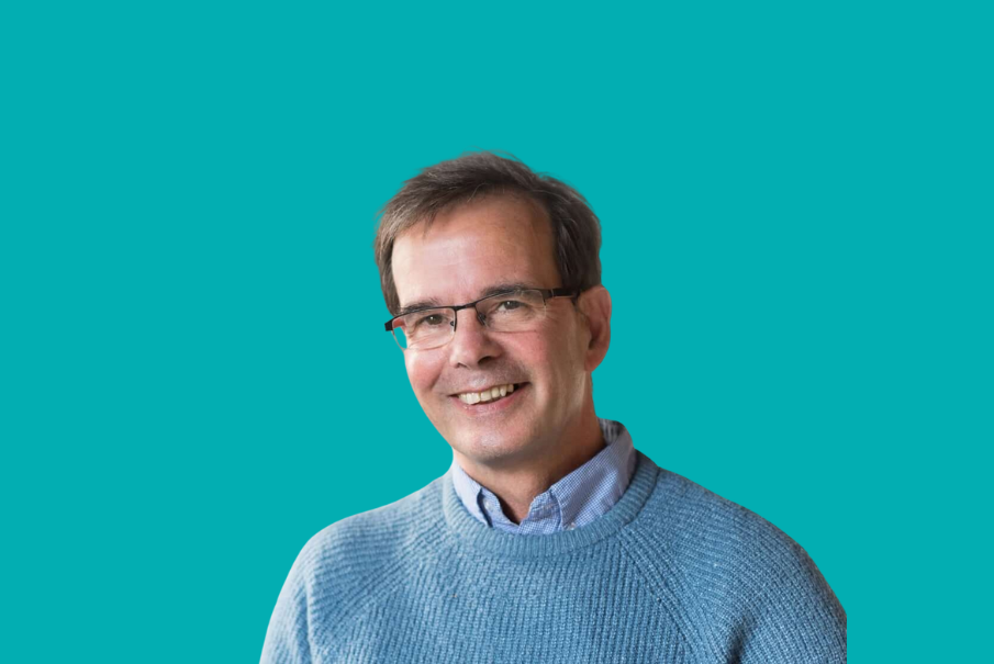 Headshot of a manin in glasses with shot dark hair, smiling. He is wearing a jumper with a shirt.