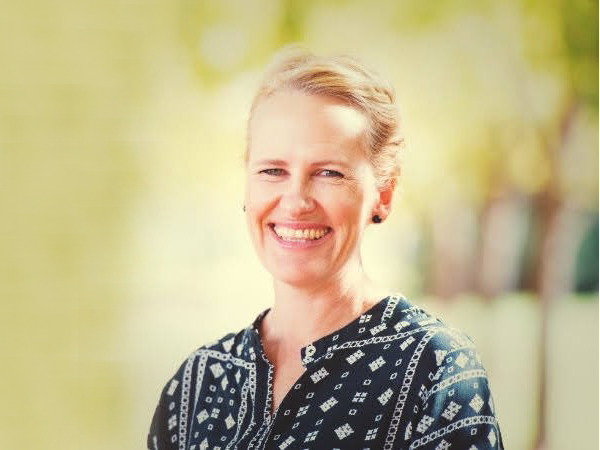 Head and shoulders image of a woman with tied back blonde hair, she is wearing a dark patterned blouse and earrings, she is smiling.