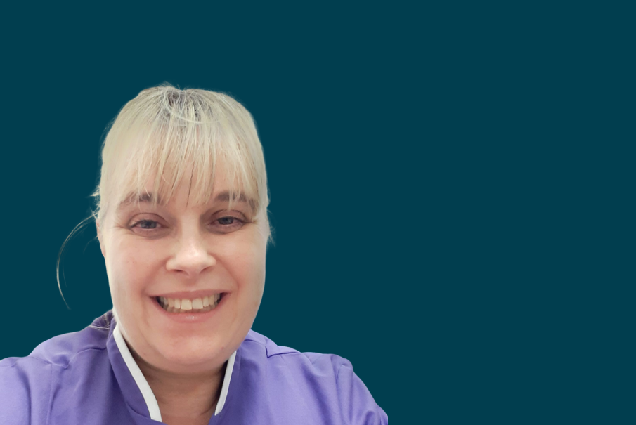 Headshot of woman in a lilac nursing uniform, with blode hair tired back and a fringe. She is smiling.