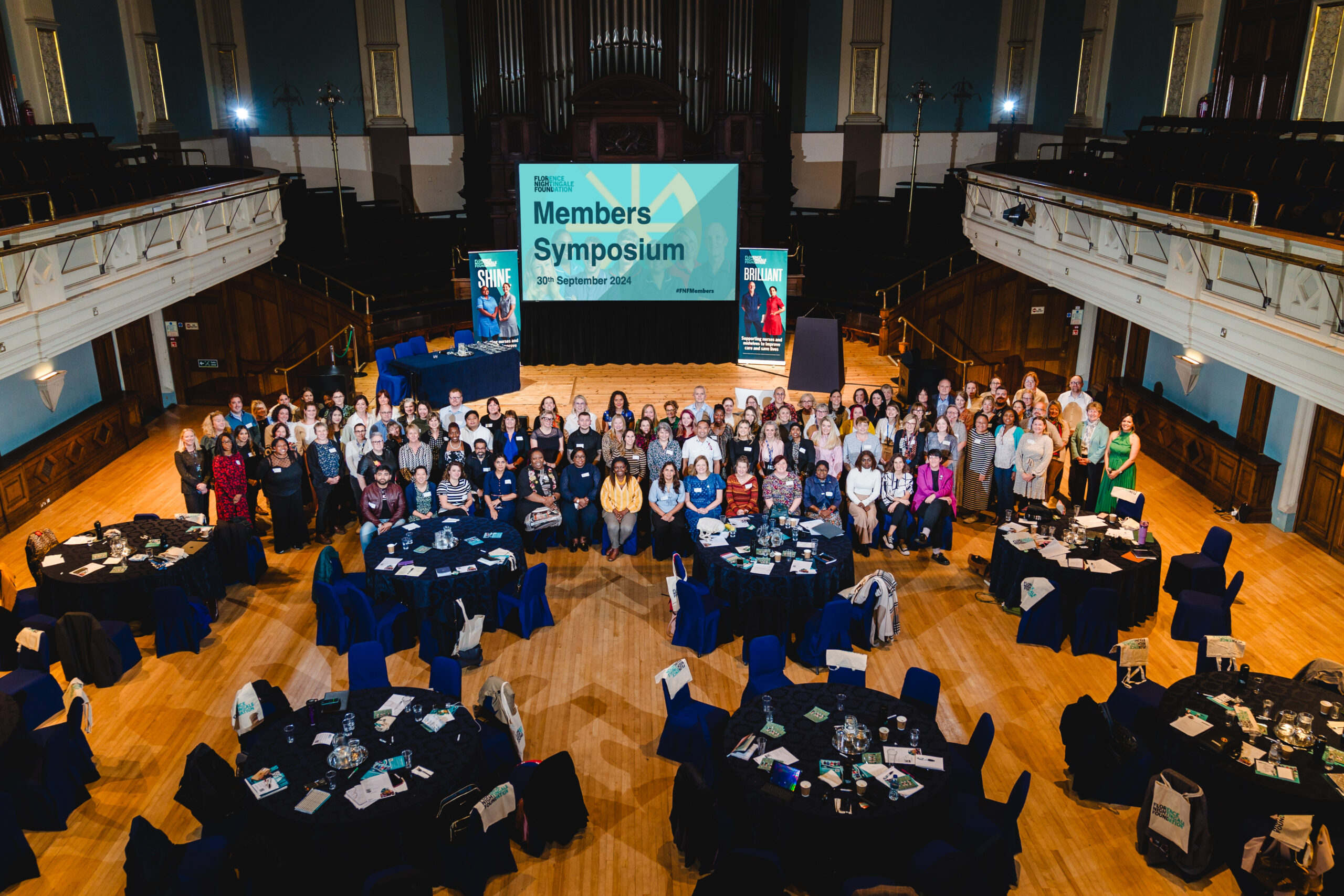 An image looking down on a large group of people smiling up at the camera, they are standing behind round tables and there is a screen in the background which says Members Symposium.