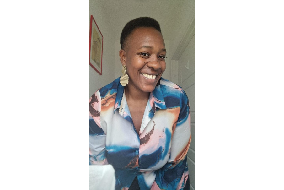 Head and shoulders image of a woman with short balck hair, She is wearing a pink, blue and white blouse and she is smiling. She is wearing earrings.