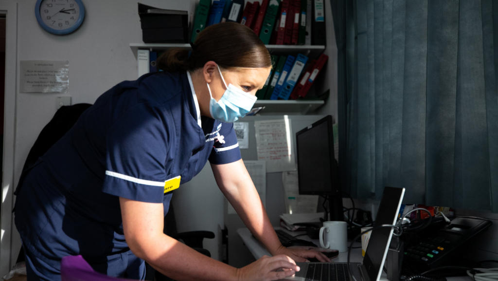 A nurse leaning over a desk, she is wearing a light blue facemask and is using a laptop.