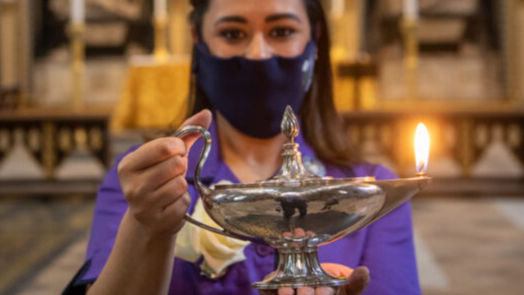 Nurse in a purple uniform, wearing a black facemask and holding a lighted lamp which is silver.