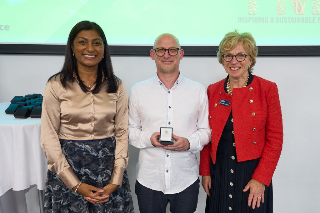 Three people standing in a row, all smiling. A woman on the left with long dark hair and a gold blouse, the middle is a man with glasses and facial hair, he is holding a box with a badge in it. The woman on the right is wearing a red jacket and a black dress and is wearing glasses.