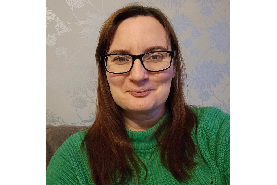 Headshot of a woman smiling. She has long dark brown hair and is wearing glasses. She is wearing a green jumper.