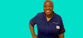 An image of a woman in a dark blue nursing uniform, she has her right hand on her right hip, she is smiling. She has very short grey hair.