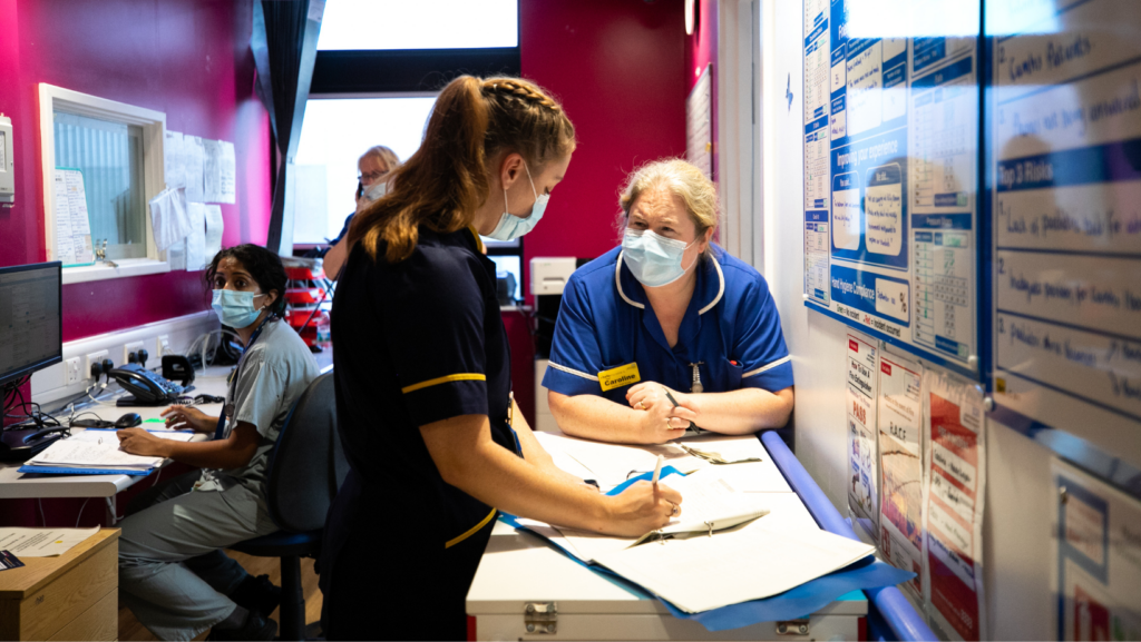 Image of two nurses in a buy office they are wearing a dark blue and a lighter blue uniform and are wearing face masks.