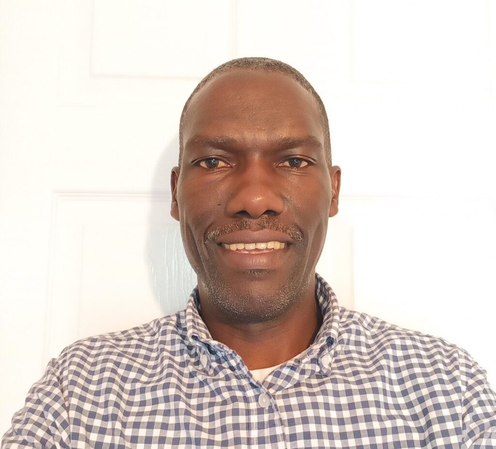Head and shoulders of a man with very short hair, wearing a checked shirt smiling.