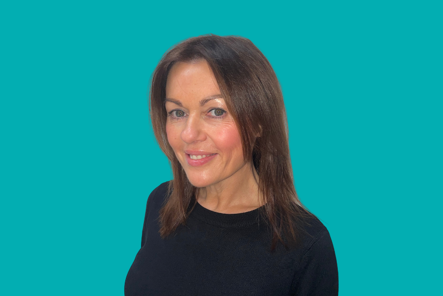 Head and shoulders image of a woman on a teal background. She has long dark hair parted in the centre, and is wearing a dark towp. She is smiling.