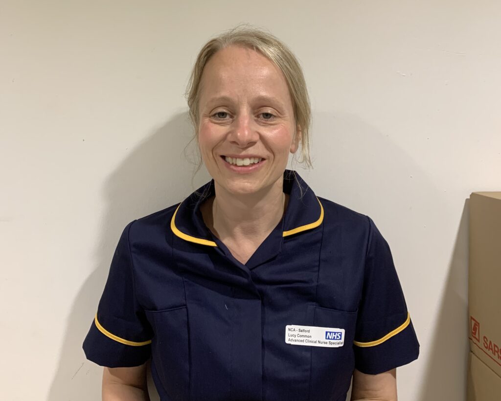 A woamn with blond hair tied back, she is wearing a dark blue uniform with yellow edges and a name badge, she is smiling.