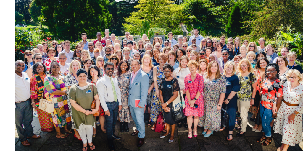An image of a large group of people standing and smiling in the sunshine, they are outside with trees and shrubbery in the background.