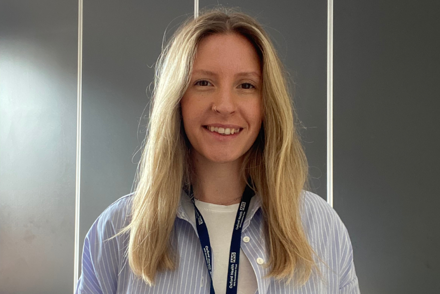 A head and shoulders image of a woman with long blong hair, she is wearing a striped blue and white shirt, with a whit tshirt, and has an NHS blue lanyard. She is smiling. There is a dark grey background.