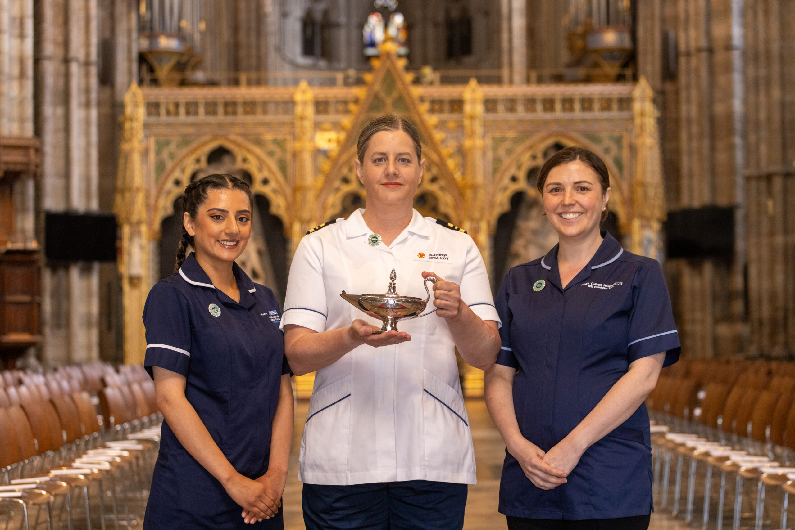 Three women in nurses uniform, standing in Westminster Abbey. The person in the middle is wearing a white uniform and dark trousers, the women to the sides are wearing dark nacy blue uniforms. The woman in the middle is holding a lamp.