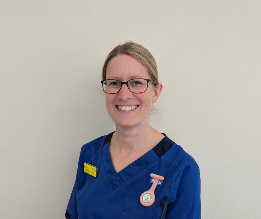 Head and shoulders image of a woman in a mid blue nursing uniform. She has a yellow name badge on the left of her uniform. She is wearing glasses and smiling. She has tied back blonde hair.