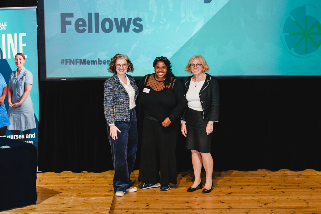 An image of three women standing on a stage with a large screen behind which says Fellows.