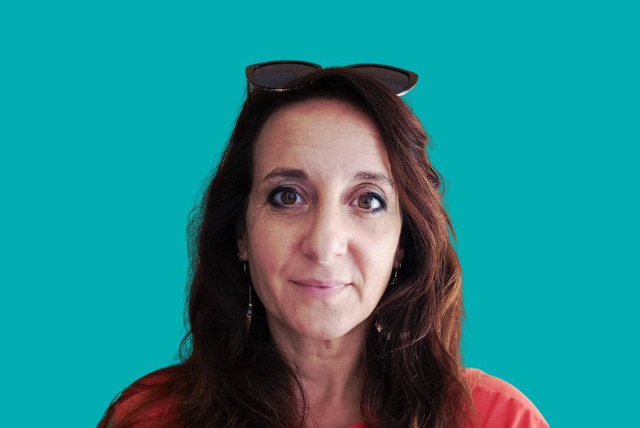 A head and shoulders image of a woman with long dark hair in an red/orange top. She has sunglasses on the top of her head and there is a teal coloured background to the image.