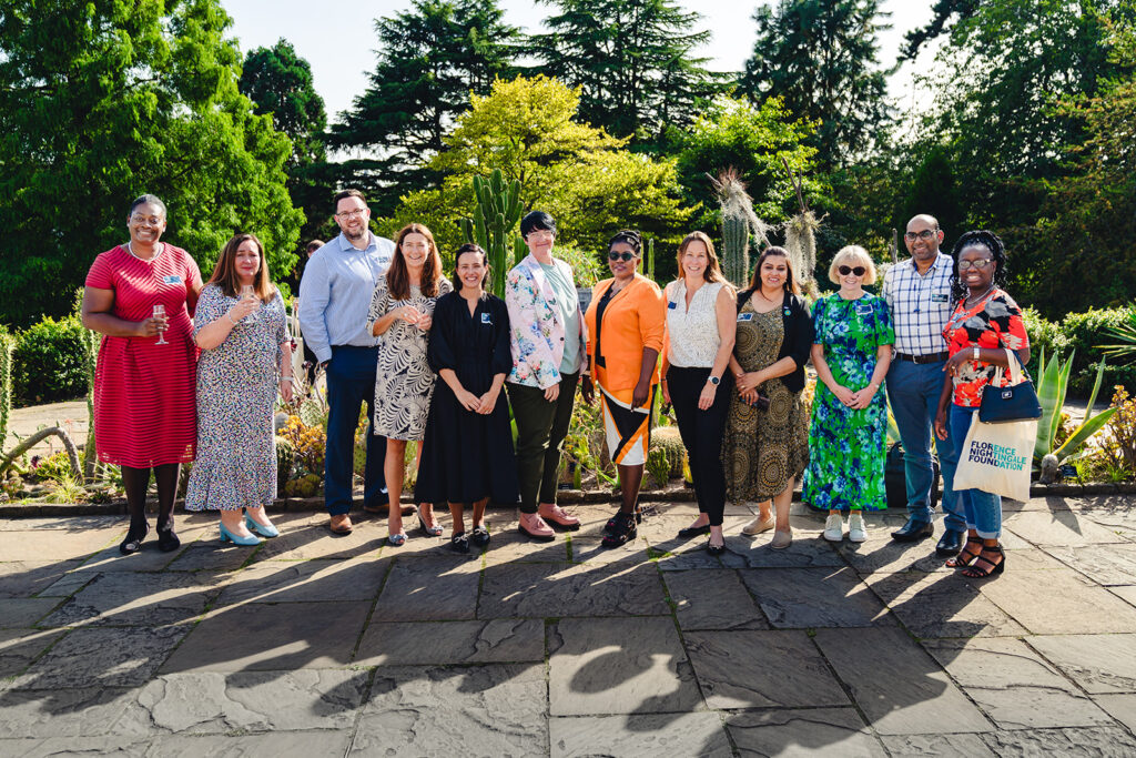 Group of people standing outside in the sunshine, smiling, there are trees in the background.