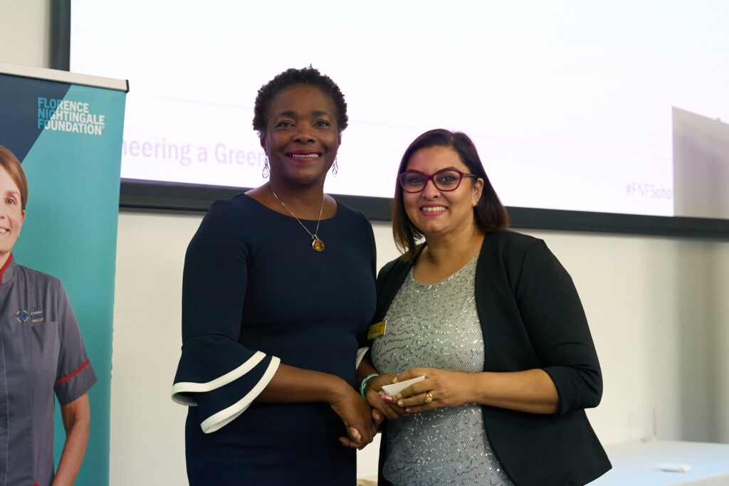 Two women standing smiling, the one on the left has a dark dress on with white edging on the sleeve, she is wearing a pendant and earrings and has short dark hair. she is holding the hands of the woman on the right who is wearing glasses. She has shoulder length dark hair and is wearing a black jacket and a sparkly silver top.