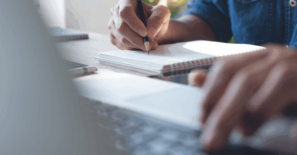 An image of the edge of a laptop with someone pressing a key, in the background there is a hand holding a pencil and writing on a notepad. This is a close up image.