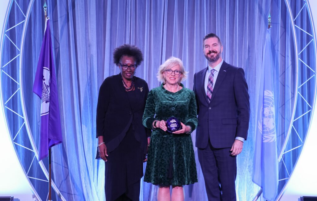 Image of three people standing. A woman with dark hair in a black suit on the left, a woman with blonde hair and glasses holding an award is wearing a green dress and on her left a man with dark hair and a beard wearing a blue suit.