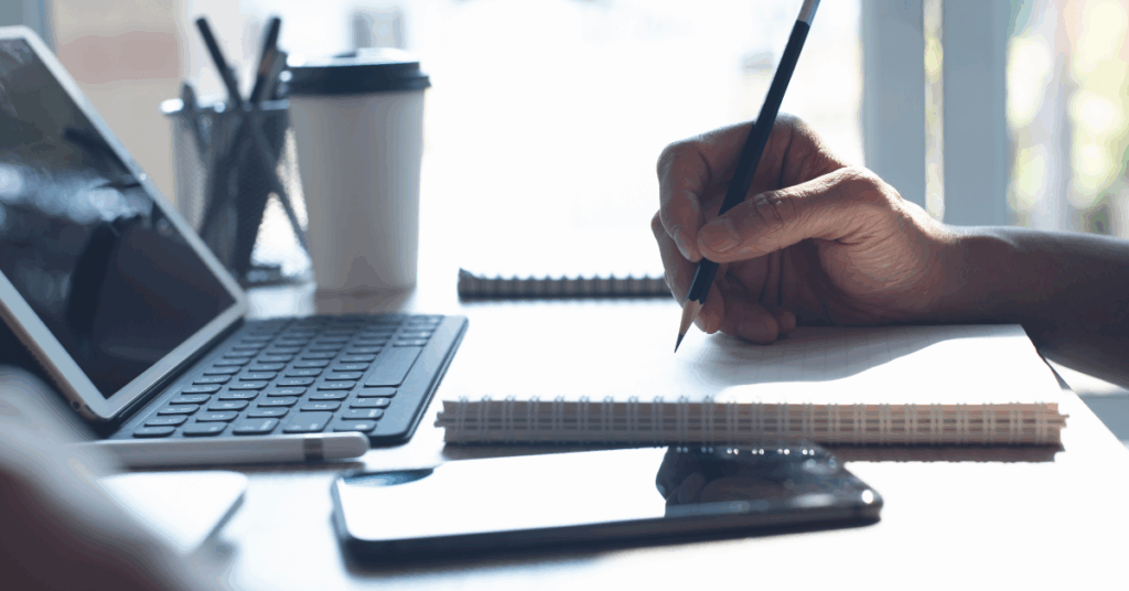 Side image of a hand on a desk holding a pencil. There is a laptop, a tall drinking cup and the hand is writing on a notepad. There is a phone at the front of the image.