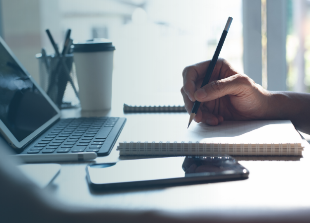 Side image of a hand on a desk holding a pencil. There is a laptop, a tall drinking cup and the hand is writing on a notepad. There is a phone at the front of the image.