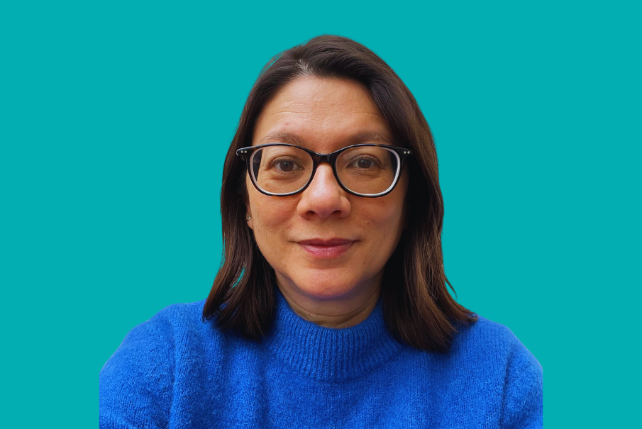 Head and shoulders image of a woman with shoulder length dark hair, she is smiling and wearing dark glasses and a royal blue jumper.
