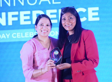 Two women with dark hair, smiling, they are holding an award together made of glass. The woman on the left is wearing pink, the woman on the right is wearing a red jacket.