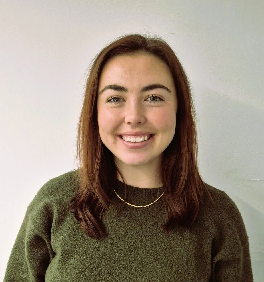 Head and shoulders image of a woman with long brown hais she is smiling and wearing an olive coloured jumper.