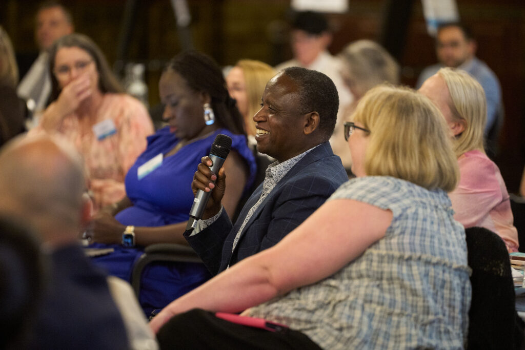 A side view image of people sitting at tables, a man is sat speaking into a handheld microphone and the audience is listening.
